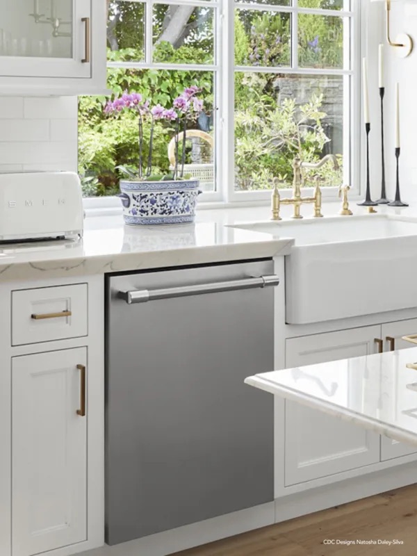 Stainless-steel dishwasher front set into white shaker cabinetry beside a farmhouse sink, orchids on the marble counter